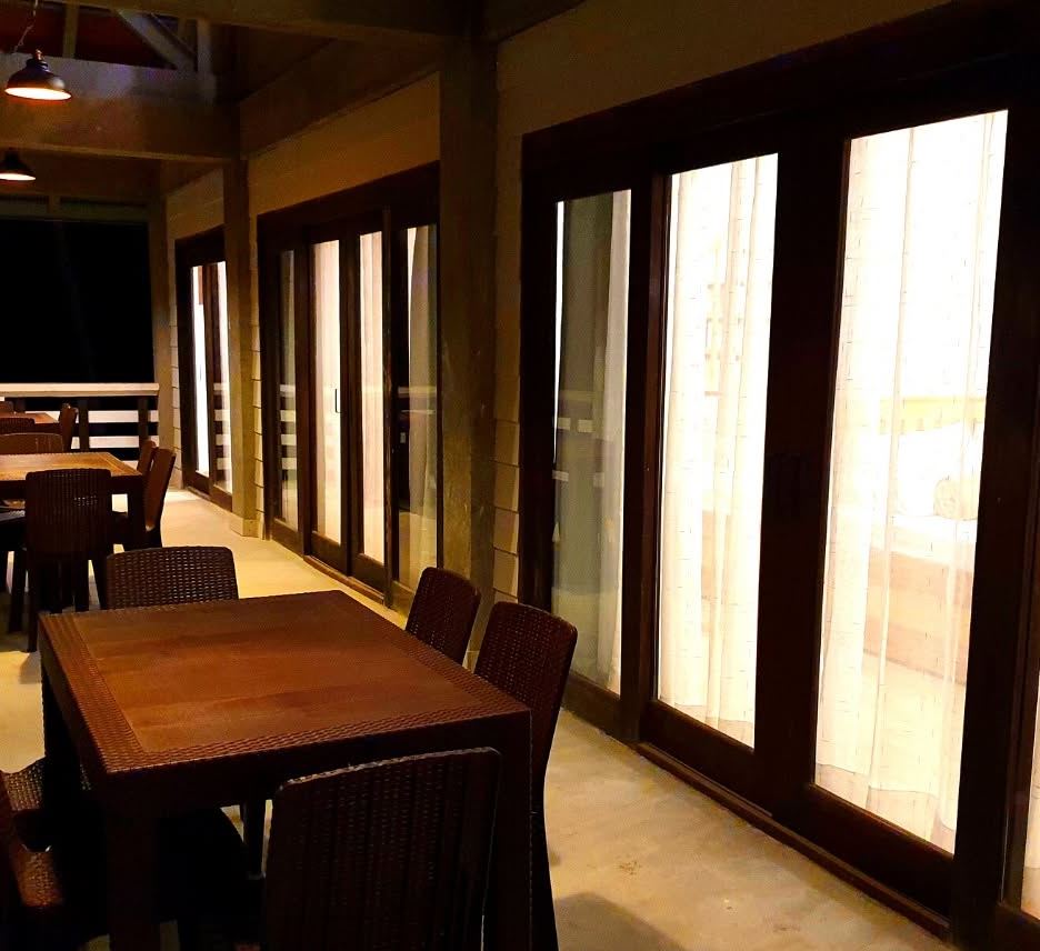 Dark timber dining tables and chairs along tall glass doors on the café veranda, warmly lit in the evening