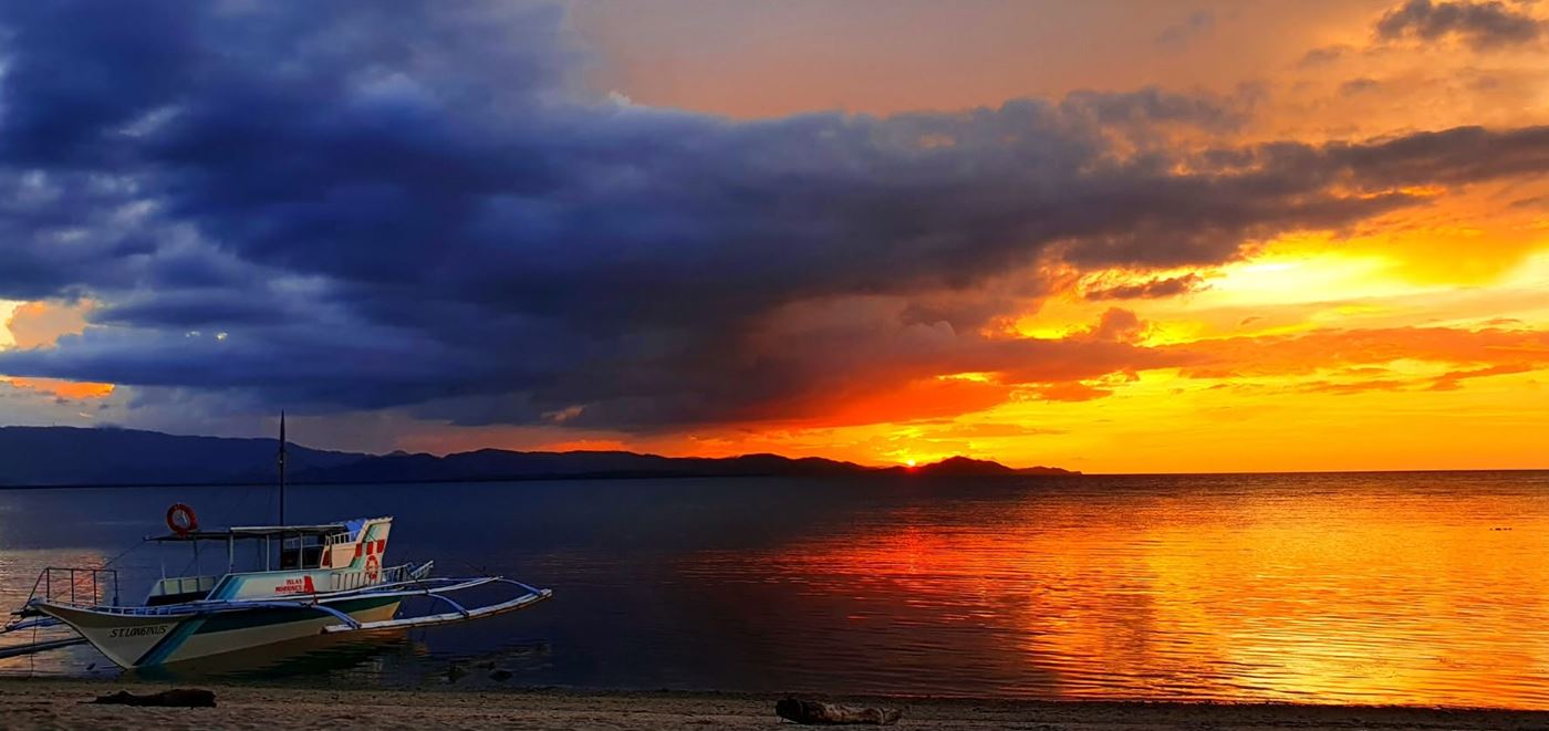An outrigger boat at the shore beneath an orange sunset over the water